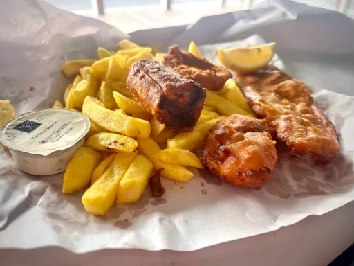 A fish and chips meal on an open paper wrapping featuring a small container of tartare sauce, a deep fried dim sim, a calamari ring, and a battered scallop. The chips and fried fish fillet have a wedge of lemon and also a sprinkling of chicken salt. 